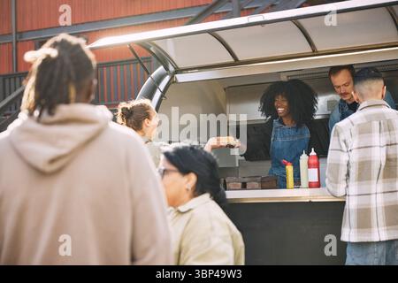 Glückliche Frau mit lockigen Haaren, die dem Gast im Food Truck einen Hot Dog zum Mitnehmen gibt Stockfoto