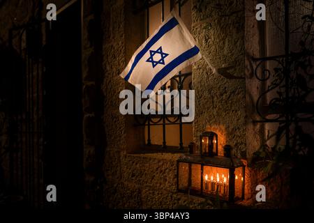 Eine israelische Flagge, die an einem Steingebäude befestigt ist, fliegt über einer Hanukka-Menora, die während des Lichterfestes mit brennenden Ölkerzen und Flammen leuchtet Stockfoto