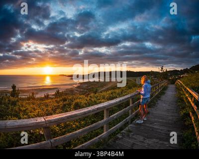 Frau steht auf einem Holzsteg über der Atlantikküste in Galicien, Spanien, und beobachtet den dramatischen Sonnenuntergang über Strand und Dünen. Die Szene vermittelt Frieden, Achtsamkeit Stockfoto