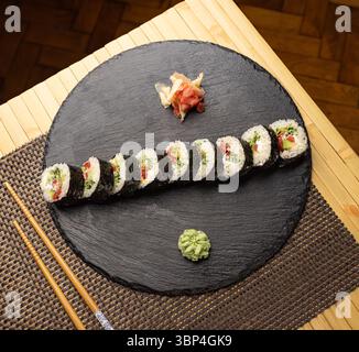 Frisch zubereitete vegane Sushi-Brötchen, gefüllt mit buntem Gemüse, serviert auf einem schwarzen Schieferteller mit Wasabi und eingelegtem Ingwer Stockfoto