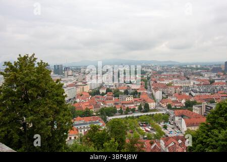 Ein weitläufiger Blick auf die Höhe fängt das Stadtbild von Ljubljana, Slowenien, an einem bewölkten Tag ein. Rot gekachelte Dächer dominieren den Vordergrund und zeigen die Stockfoto