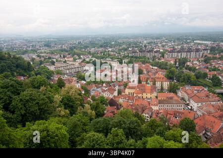 Ein weitläufiger Blick auf die Höhe fängt das Stadtbild von Ljubljana, Slowenien, an einem bewölkten Tag ein. Rot gekachelte Dächer dominieren den Vordergrund und zeigen die Stockfoto