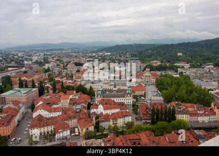 Ein weitläufiger Blick auf die Höhe fängt das Stadtbild von Ljubljana, Slowenien, an einem bewölkten Tag ein. Rot gekachelte Dächer dominieren den Vordergrund und zeigen die Stockfoto