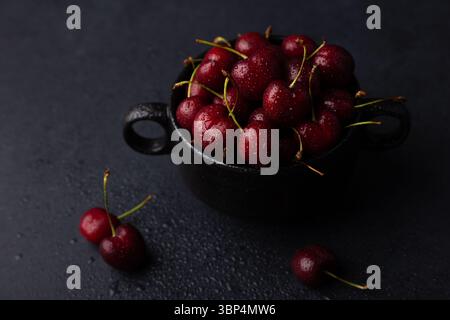 Saftige, reife frische Kirschen mit Wassertropfen auf einem schwarzen Teller auf schwarzem Hintergrund. Saisonale Früchte, Ernte. Selektiver Fokus, Nahaufnahme. Stockfoto