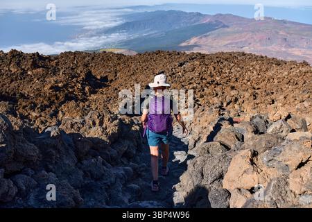 Frau, die den felsigen vulkanischen Pfad vom Gipfel des Pico del Teide in Richtung La Rambleta mit bewaldeten Hängen und Küste in der Ferne absteigt Stockfoto