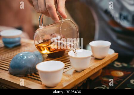 Geben Sie während der Zeremonie goldenen Tee von Hand aus klarem Pitcher in eine weiße Tasse auf Holztablett. Nahaufnahme des eleganten Teerituals. Stockfoto
