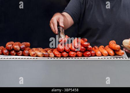 Man kippt und stapelt heiße gegrillte Würstchen auf einer Metalltheke, serviert Kunden auf dem Markt, Street Food und Barbecue Stockfoto