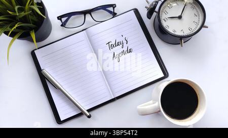 „Today's Agenda“ Notizbuch, Kaffee, Wecker und Brille auf weißem Hintergrund Stockfoto