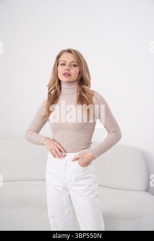 Elegante Frau mit langen Haaren, mit beigefarbenem Rollkragen und weißer Hose, steht selbstbewusst vor einem schlichten Hintergrund. Ihre Hände liegen in Taschen Stockfoto
