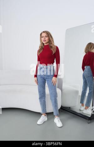 Eine Frau in rotem Pullover und blauer Jeans steht selbstbewusst neben einem großen Bodenspiegel, der ihr Bild reflektiert, in einem modernen, minimalistischen Wohnbereich mit elegantem Design Stockfoto