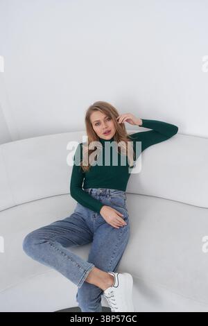 Die Frau sitzt auf einem geschwungenen weißen Sofa, trägt einen grünen Ripppullover und blaue Jeans. Ihr rechter Arm liegt auf dem oberen Rand, während ihre linke Hand in ihr ist Stockfoto