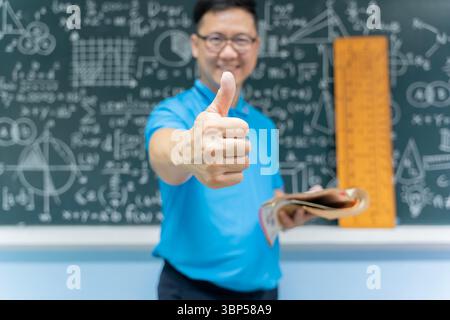 Glücklicher, selbstbewusster Lehrer im Klassenzimmer, der ein Buch hält und die Daumen nach oben zeigt. Er steht mit mathematischen und naturwissenschaftlichen Formeln vor der Tafel Stockfoto