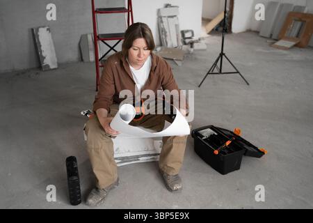 Reparaturfrau liest Papierpläne, während sie mit Toolbox und Spirit Level in einem nicht renovierten Zimmer sitzt Stockfoto