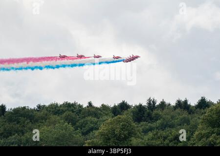 Rote Pfeile fliegen in Formation über dichten grünen Wäldern neben Silverstone und lassen rote und blaue Rauchpfade unter einem hellen bewölkten Himmel frei Stockfoto