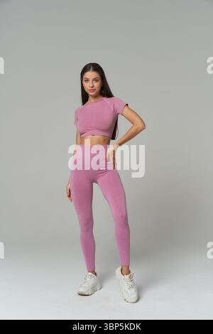 Eine junge Frau in rosa, sportlichem Outfit und weißen Sneakers zeigt eine selbstbewusste Pose. Ihr langes Haar ist unten und sie steht mit einer Hand auf der Hüfte Stockfoto