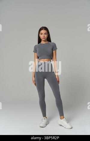 Eine Frau steht selbstbewusst in einem passenden grauen Crop-Top und Leggings mit weißen Sneakern. Ihr langes, glattes Haar und ihr neutraler Ausdruck vermitteln ein Stockfoto