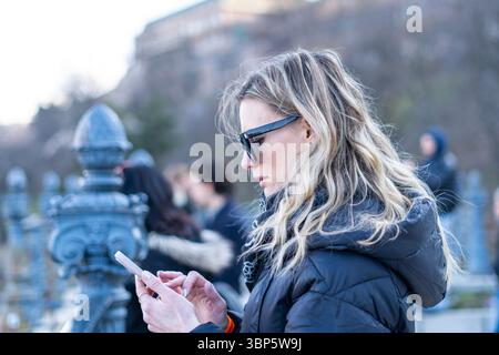 Blonde Frau in schwarzem Mantel und Sonnenbrille mit ihrem Smartphone im Winterpark Stockfoto