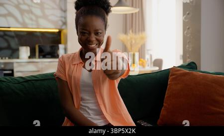 Glückliche fröhliche lächelnde afroamerikanische Geschäftsfrau weibliche Geschäftsfrau Mädchen Hausbesitzer sitzende Couch Sofa Blick in die Kamera zeigt Daumen nach oben Geste Stockfoto