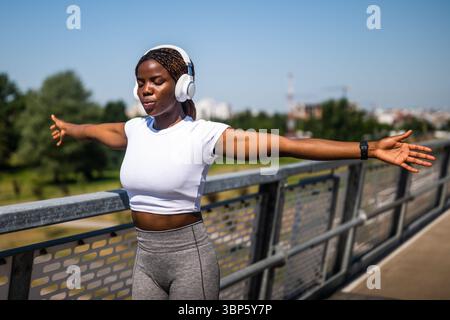 Afroamerikanische Frau in Sportbekleidung, die Sport- und Stretchanwendungen trägt und Musik über kabellose Kopfhörer auf der Brücke in der Stadt hört. Stockfoto