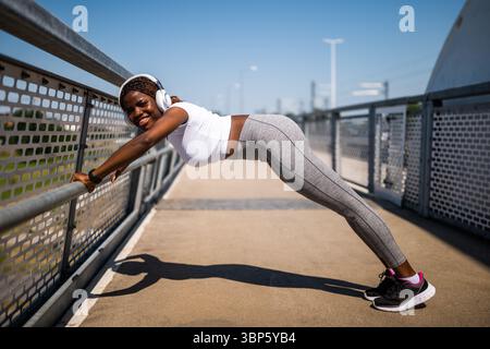 Afroamerikanische Frau in Sportbekleidung, die Liegestütze macht und Sport macht, während sie Musik über Kopfhörer auf der Stadtbrücke hört. Stockfoto