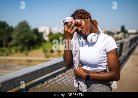 Afroamerikanische Frau, die enttäuscht und besorgt aussah, während sie nach intensivem Training auf der Stadtbrücke mit einem Handtuch den Schweiß vom Gesicht abwischte. Stockfoto