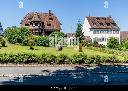 Fachwerkhaus mit rundem Erkerfenster direkt gegenüber St. Maria und Markusminster in Mittelzell. Das zweigeschossige Fachwerkgebäude mit polygonalem Erkerfenster ist mit dekorativen Fachwerkelementen wie Andreaskreuzen versehen, die typisch für die alemannische Tradition sind. Blick auf traditionelle Häuser und gepflegte Gärten am Münsterplatz auf der Insel Reichenau. Die Szene zeigt die idyllische Atmosphäre dieses UNESCO-Weltkulturerbes unter einem blauen Himmel. Münsterplatz, Reichenau, Baden-Württemberg, Deutschland Stockfoto