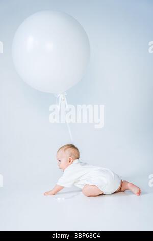 Baby krabbelt auf dem Boden mit großem weißen Ballon auf hellem Hintergrund. Stockfoto