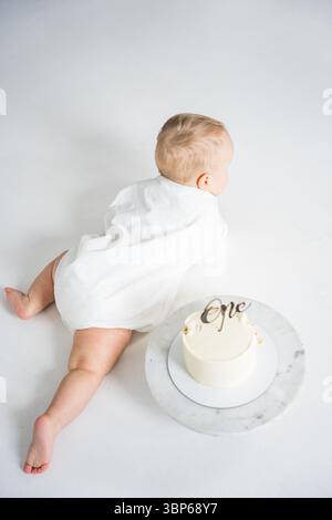 Baby krabbelt in der Nähe der ersten Geburtstagskuche auf weißem Boden Hintergrund. Stockfoto