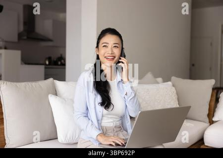 Frau, die auf dem Sofa sitzt und mit Laptop Gespräche auf dem Smartphone führt Stockfoto