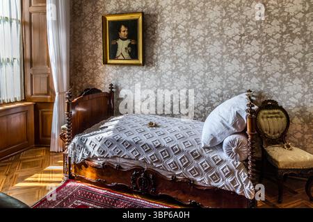 Das historische Schlafzimmer von Louis Napoleon auf Schloss Arenenberg in Salenstein, Thurgau, Schweiz, ist auf dem Bild zu sehen. Das einfache Zimmer ist mit einem antiken Holzbett, einer weißen Bettdecke und einem kunstvollen Stuhl ausgestattet. An der Wand hängt ein Porträt von Napoleon Bonaparte, das die enge Verbindung der Burg zur Familie Bonaparte unterstreicht, da es die ehemalige Residenz von Hortense de Beauharnais war, Napoleons Stieftochter. Das Bild fängt die authentische historische Atmosphäre dieses gut erhaltenen Zimmers ein Stockfoto
