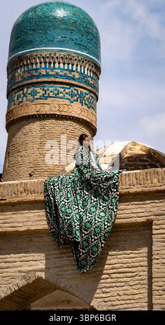 Eine Frau in einem fließenden grün-weiß gemusterten Kleid sitzt anmutig auf einem historischen Gebäude mit einer blauen Kuppel. Die Architektur zeichnet sich durch eine komplizierte Gestaltung aus Stockfoto