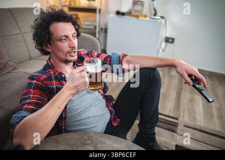 Ein junger Mann genießt ein Bier, während er in seinem Wohnzimmer Fernsehen sieht Stockfoto
