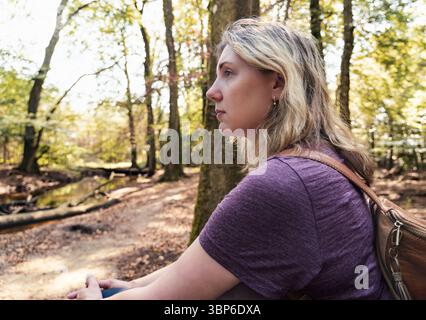 Eine Frau genießt einen Moment, während sie allein auf einem Waldweg sitzt, umgeben von hohen Bäumen und verflecktem Sonnenlicht. Das Schweigen lässt tiefe Gedanken zu Stockfoto