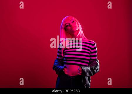 Auffällige, stilvolle Frau mit rosa Haaren präsentiert sich im Studio über isoliertem Hintergrund. Kreatives weibliches Modell zeigt einen kantigen Look bei selbstbewussten Fotoshootings unter dramatischer pinkfarbener Beleuchtung. Stockfoto