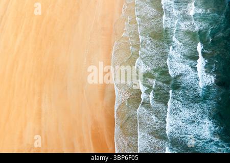 Von oben aus blickt man auf einen sonnigen Sandstrand mit sanften weißen, schaumigen Wellen Stockfoto