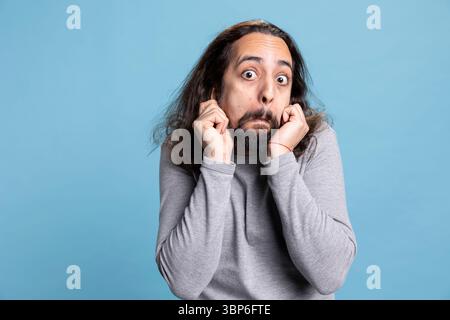 Fröhlicher bärtiger Mann mit langen lockigen Haaren schlägt eine alberne Pose und zeigt eine sorglose Haltung. Spielerisches Verhalten und komisches Gesicht unterstreichen seine humorvolle Persönlichkeit in diesem Studio vor blauem Hintergrund. Stockfoto
