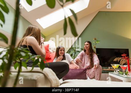 Drei Frauen sitzen auf einer bequemen Couch in einem warm beleuchteten, mit Pflanzen dekorierten Raum und genießen die Gesellschaft der anderen, umgeben von positiver Energie und Mo Stockfoto