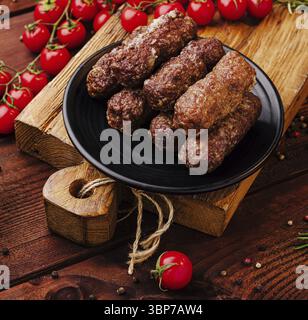 Essen, dicke Fleischbrötchen sind auf einem schwarzen Teller auf einem Holzbrett angeordnet, umgeben von frischen Kirschtomaten und verschiedenen bunten Gewürzen, Crea Stockfoto
