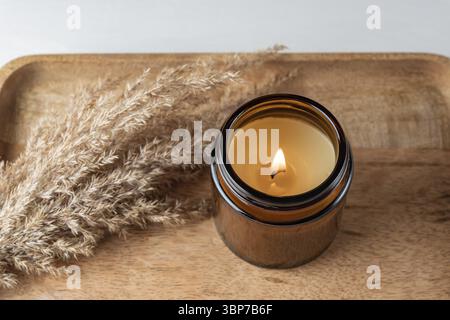 Handgemachte Kerze aus Paraffin und Sojawachs im Glasgefäß mit trockenem Schilfgras auf Holztablett. Kerzenherstellung Stockfoto