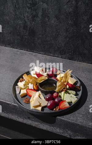 Speisen, Ein Gourmet-Arrangement umfasst verschiedene Käsesorten, frische Erdbeeren, Trauben und getrocknete Früchte auf einer schicken schwarzen Platte. Eine kleine Schüssel Honig liegt Stockfoto