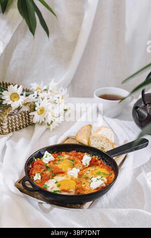 Essen, Shakshuka mit Eiern, Fetakäse und Kräutern serviert in einer gusseisernen Pfanne auf einer weißen Tischdecke mit einer Tasse Tee, ein gesundes und köstliches Frühstück Stockfoto