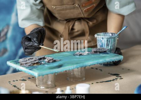 Die Künstlerin gießt Epoxidharz und verschmiert es mit einem Stock auf Holzbrett. Handwerker mit flüssiger Kunsttechnik, die ein Bild von Meeresschaum und Welle erstellt Stockfoto