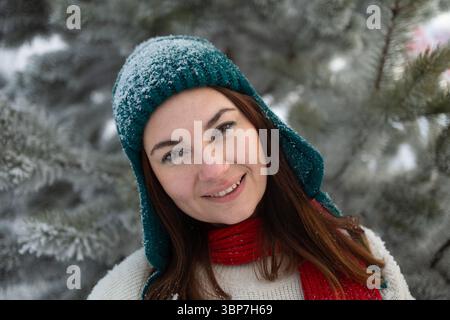 Eine fröhliche Frau, die einen bunten Strickmütze und einen gemütlichen Schal trägt, lächelt in einer malerischen, verschneiten Winterlandschaft Stockfoto