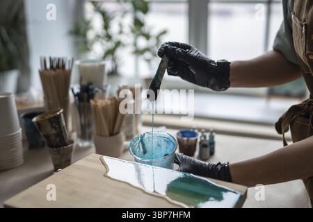 Die Künstlerin gießt Epoxidharz auf Holzbretter. Handwerkerin mit flüssiger Kunsttechnik, das Bild von Meer und Wellen auf Leinwand, Nahaufnahme. Ar Stockfoto