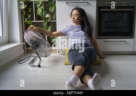 Überhitzung in Haushalten. Müde überhitzte junge asiatische Frau, die auf dem Boden in der Küche in der Nähe eines elektrischen Ventilators saß und sich zu Hause während der extremen Sommerhitze abkühlte Stockfoto