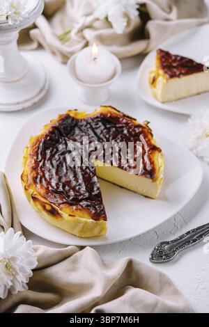 Essen, baskischer Gourmet-Käsekuchen mit einer Scheibe serviert auf weißem Teller, begleitet von zündeten Kerzen und Blumen Stockfoto
