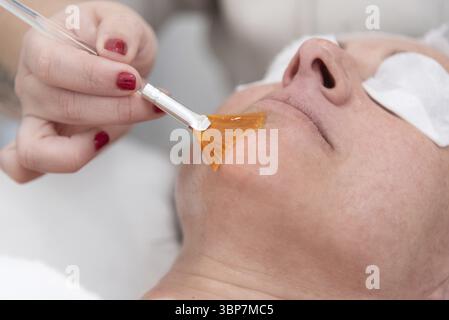 Kosmetikerin, die eine orangene Kosmetikmaske mit einem Pinsel auf das Gesicht einer Frau aufträgt, während sie sich in einem Schönheitssalon entspannt und sich mit verjüngender Pflege verwöhnt Stockfoto