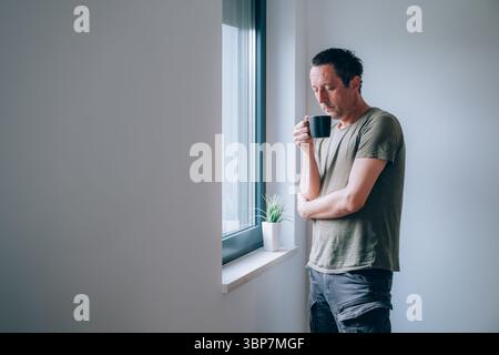 Nachdenklicher Mann, der morgens Kaffee am Fenster der Wohnung trinkt, während er hinausschaut und nachdenkt. Coffeindosis zum Tagesbeginn. Selektiv fo Stockfoto