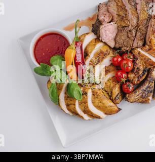 Essen, Blick über dem Kopf auf köstliche gegrillte Fleischscheiben, serviert mit frischem Gemüse und Dippsoße auf einem weißen Teller Stockfoto