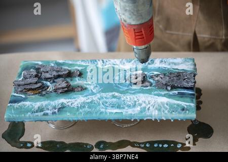 Künstler arbeitet mit flüssigem Epoxidharz, erzeugt Meer, Wellen und Felsen auf zweiteiligen Harzplatten, bläst ein Holzgemälde mit der Konstruktion thermisch trocken Stockfoto